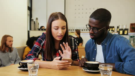 Caucasian-woman-and-african-american-man-watching-a-video-on-smartphone-in-a-cafe