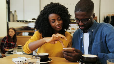 African-american-man-and-woman-watching-a-video-on-smartphone-sitting-at-a-table-in-a-cafe