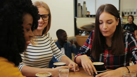 Close-up-view-of-african-american-and-caucasian-women-friends-laughing-and-watching-something-on-a-smartphone-sitting-at-a-table-in-a-cafe