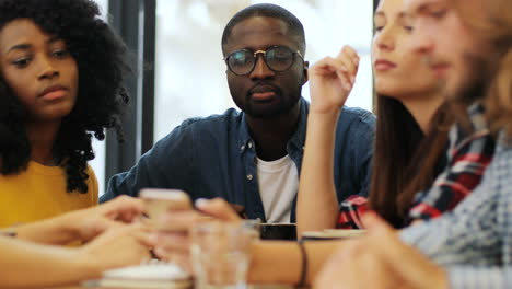 Close-up-view-of-multiethnic-group-of-friends-talking-and-using-smartphone-sitting-at-a-table-in-a-cafe