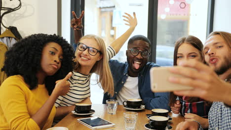 Close-up-view-of-multiethnic-group-of-friends-making-a-selfie-with-a-smartphone-and-doing-funny-gestures-sitting-at-a-table-in-a-cafe