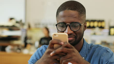 Vista-Cercana-Del-Hombre-Afroamericano-Sonriendo-Y-Enviando-Mensajes-De-Texto-En-Un-Teléfono-Inteligente-Sentado-En-Una-Mesa-En-Un-Café