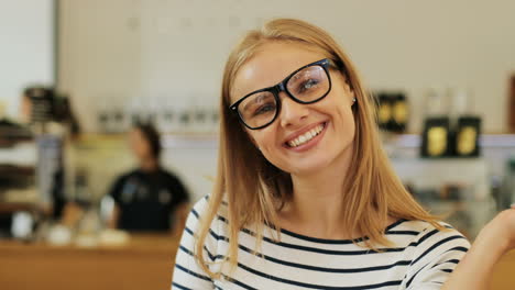 Close-up-view-of-caucasian-blonde-woman-in-glasses-smiling-at-camera-sitting-at-a-table-in-a-cafe