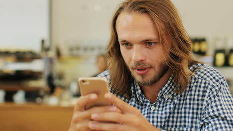 Close-up-view-of-caucasian-blond-man-with-long-hair-texting-on-the-phone-sitting-at-a-table-in-a-cafe