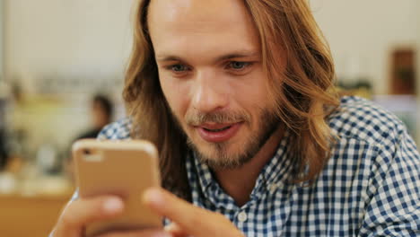 Close-up-view-of-caucasian-blond-man-with-long-hair-texting-on-the-phone-sitting-at-a-table-in-a-cafe