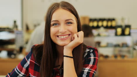 Close-up-view-of-brunette-woman-with-long-hair-smiling-at-camera-sitting-at-a-table-in-a-cafe