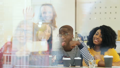 Camera-focuses-on-a-multiethnic-group-of-friends-through-the-window-making-a-selfie-and-doing-funny-gestures-while-they-are-sitting-at-a-table-in-a-cafe