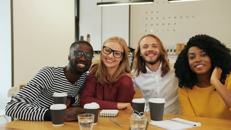 Multiethnic-group-of-friends-smiling-and-looking-at-camera-sitting-at-a-table-in-a-cafe