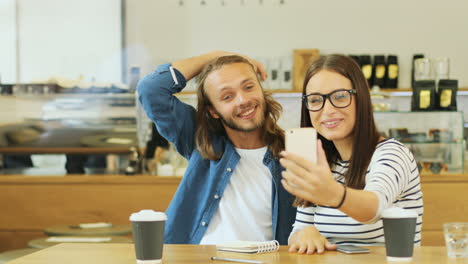 Caucasian-man-and-woman-friends-making-a-video-call-using-smartphone-sitting-at-a-table-in-a-cafe