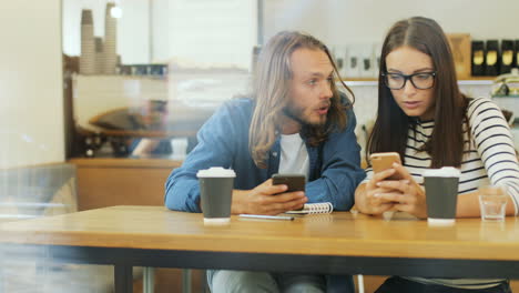 Caucasian-man-and-woman-friends-using-smartphone-and-talking-while-they-are-sitting-at-a-table-in-a-cafe