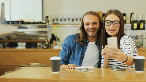 Caucasian-man-and-woman-friends-making-a-selfie-using-smartphone-sitting-at-a-table-in-a-cafe
