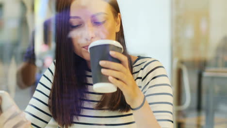 Camera-focuses-on-a-brunette-woman-through-the-window-drinking-coffee-and-texting-on-the-smartphone-while-she-is-sitting-at-a-table-in-a-cafe