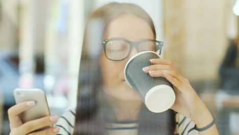 Camera-focuses-on-a-brunette-woman-in-glasses-through-the-window-drinking-coffee-and-texting-on-the-smartphone-while-she-is-sitting-at-a-table-in-a-cafe