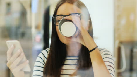 Camera-focuses-on-a-brunette-woman-in-glasses-through-the-window-drinking-coffee-and-texting-on-the-smartphone-while-she-is-sitting-at-a-table-in-a-cafe