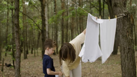 Caucasian-woman-hanging-out-washing-clothes-on-clothesline-with-the-help-of-her-son-at-the-camping-in-the-forest