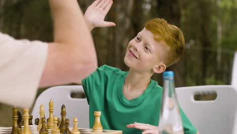 Süßer-Kleiner-Junge,-Der-Mit-Seinem-Vater-Schach-Spielt-Und-Ihm-Auf-Dem-Campingplatz-Im-Wald-High-Five-Gibt