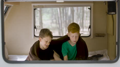 Two-little-brothers-with-blanket-on-their-shoulders-sitting-on-bed-in-a-campervan-and-reading-a-book