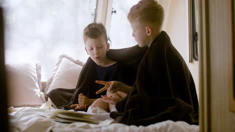 Two-little-brothers-with-blanket-on-their-shoulders-sitting-on-bed-in-a-campervan-and-playing-rock-scissors-paper