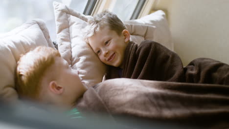 Two-little-brothers-talking-and-laughing-together-while-lying-under-a-blanket-in-the-campervan-bed