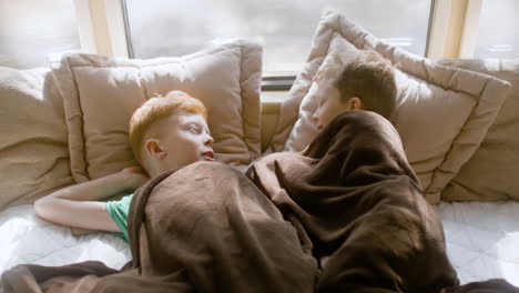 Two-little-brothers-talking-and-laughing-together-while-lying-under-a-blanket-in-the-campervan-bed