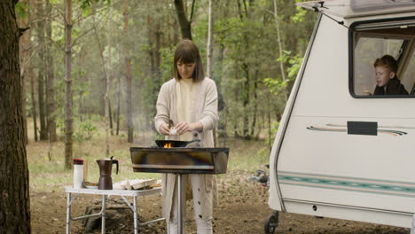 Woman-cooking-on-the-barbecue-and-breaking-eggs-in-a-pan-while-her-son-looking-out-of-campervan-window