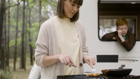Woman-cooking-sausages-on-barbecue-grill-while-her-son-looking-out-from-campervan-window