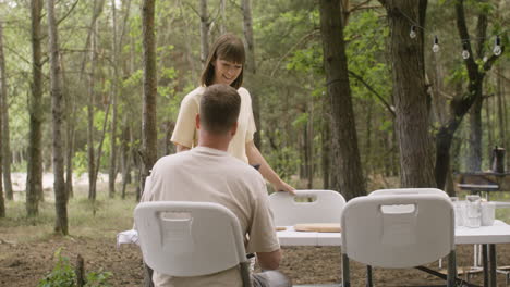Happy-woman-bringing-frying-pan-to-table-to-have-breakfast-with-her-husband-at-the-camping-in-the-forest