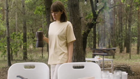 Happy-woman-bringing-coffee-pot-to-table-to-have-breakfast-with-her-husband-at-the-camping-in-the-forest