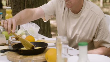 Happy-man-moving-fried-eggs-in-a-pan-with-a-wooden-fork-while-having-breakfast-with-his-family-at-the-camping-in-the-forest