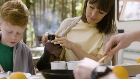 Lächelnde-Frau,-Die-Kaffee-In-Einen-Becher-Gießt,-Während-Sie-Mit-Ihrer-Familie-Beim-Camping-Im-Wald-Frühstückt