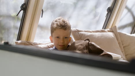 Cute-little-boy-lying-under-a-blanket-in-the-campervan-bed,-looking-at-the-camera-and-smiling