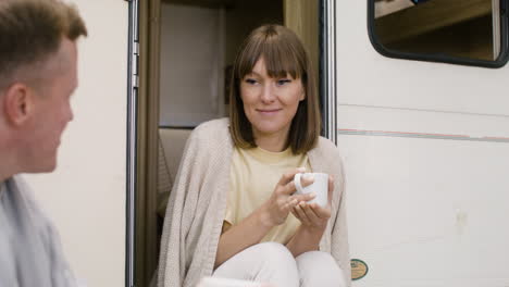 Happy-woman-sitting-on-the-stairs-of-campervan,-holding-coffee-mug-and-talking-with-man
