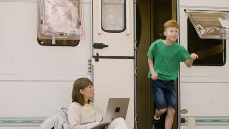 Concentrated-woman-sitting-on-chair-and-working-on-laptop-computer-at-the-camping-in-the-forest-while-her-son-getting-out-of-the-campervan