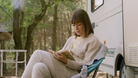 Concentrated-woman-reading-a-book-while-sitting-on-a-chair-near-a-campervan-in-the-forest