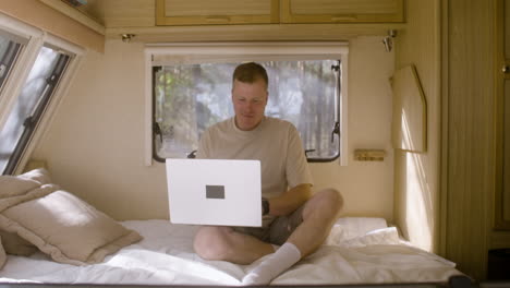 Zoom-shot-of-a-smiling-man-sitting-on-the-campervan-bed-and-working-on-laptop-computer