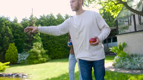 Side-view-of-caucasian-young-man-throwing-a-red-petanque-ball-in-the-park-on-a-sunny-day-while-his-friends-wait-their-turns