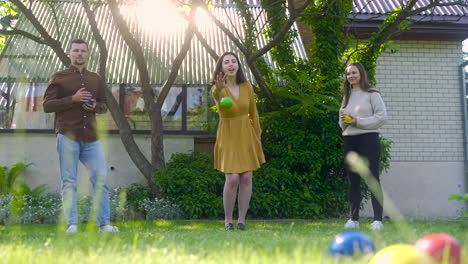Front-view-of-caucasian-young-woman-throwing-a-green-petanque-ball-in-the-park-on-a-sunny-day-while-her-friends-wait-their-turns