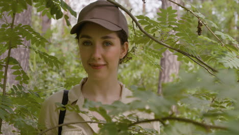 Close-up-view-of-caucasian-female-forest-warden-wearing-cap-and-looking-at-the-camera-through-the-branches-of-a-tree