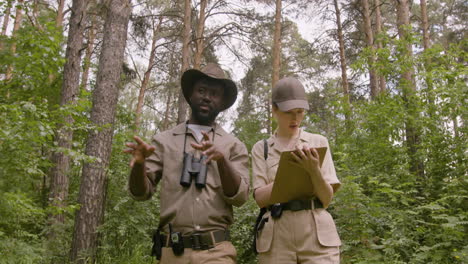 Lower-view-of-caucasian-and-african-american-forest-wardens-taking-notes-and-talking-while-walking-in-the-woods