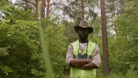 African-american-male-forest-warden-wearing-hat-and-binoculars-and-looking-at-camera-with-crossed-arms-in-the-woods