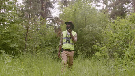 Distant-view-of-african-american-male-forest-warden-wearing-hat-and-binoculars-while-walking-and-talking-on-walkie-talkie-in-the-woods