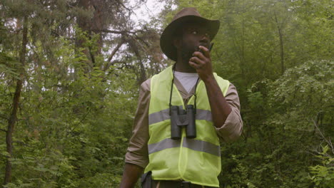 African-american-male-forest-warden-wearing-hat-and-binoculars-while-walking-and-talking-on-walkie-talkie-in-the-woods