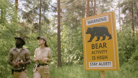 Close-up-view-of-a-signboard-to-warn-of-high-bear-activity-in-the-woods-and-two-rangers-walking-near-it