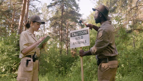 African-american-forest-warden-installing-a-signboard-to-warn-that-there-are-wild-animals-in-the-woods-while-his-female-coworker-using-a-tablet