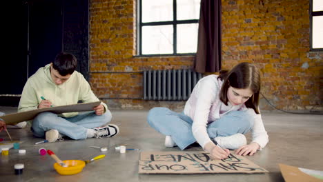 Zoom-out-of-young-environmental-activists-painting-placards-sitting-on-the-floor