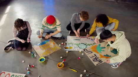 Top-view-of-young-environmental-activists-painting-placards-sitting-on-the-floor