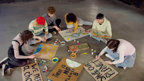 Top-view-of-young-environmental-activists-painting-placards-sitting-on-the-floor