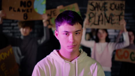 Portrait-of-a-serious-young-male-environmental-activist-looking-at-the-camera-while-his-friends-holding-placards-and-protesting-behind-him