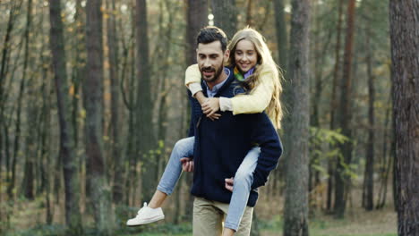 Caucasian-father-walking-in-the-forest-and-carrying-his-cute-small-daughter-on-his-back-while-they-talking