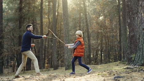 Cheerful-Caucasian-father-and-little-son-playing-in-the-forest-with-sticks-like-they-fighting-with-swords
